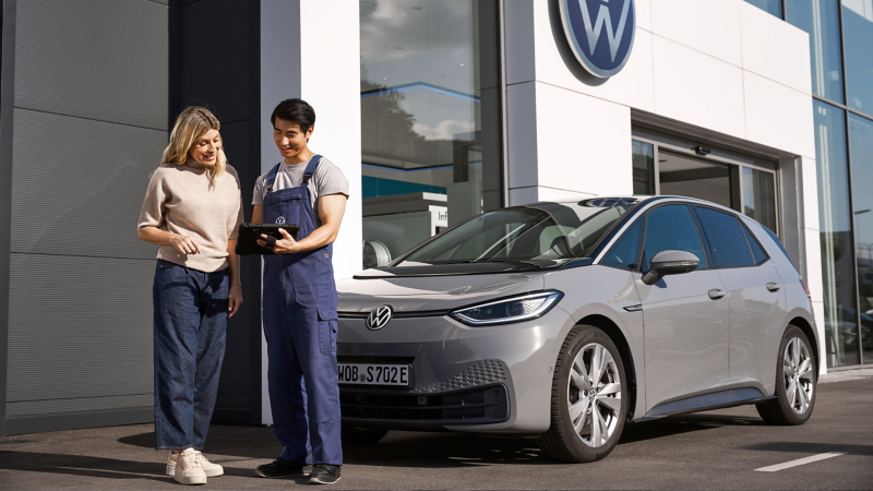 A VW service employee with a tablet in his hand in front of two VW cars next to a workshop