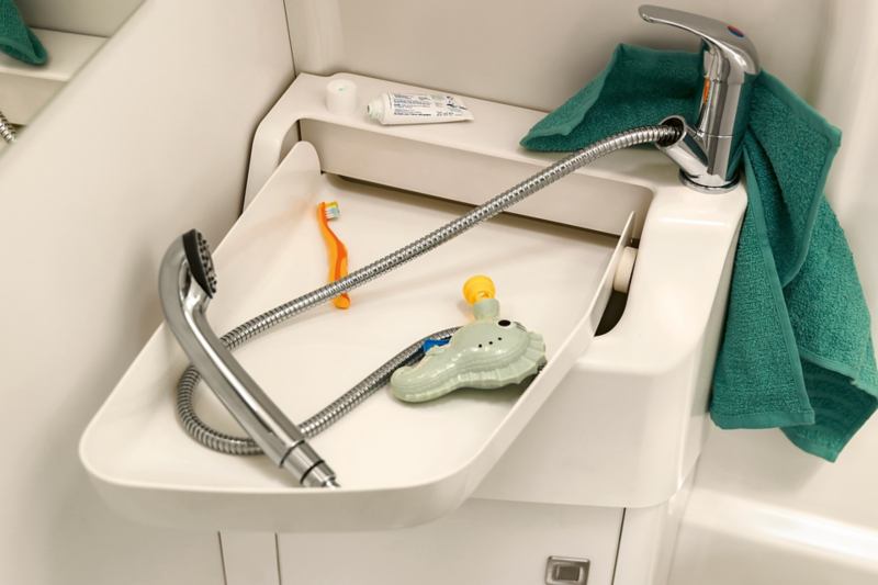 Vue en détail de la salle de bains du Grand California de Volkswagen Véhicules Utilitaires. Le lavabo flexible est déplié. Dessus se trouvent le pommeau de douche, une brosse à dents d’enfant et un jouet coloré.