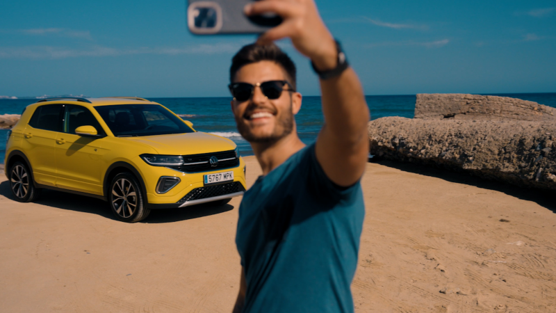 Un chico haciendose un selfie con un Volkswagen T-Cross de la gama SUV de color amarillo estacionado en la calle al lado del mar